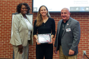 Woman poses in between two adults holding award in front of red brick background.