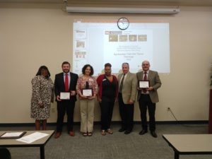 Group of adults pose displaying awards in their hands. They are standing in front of a powerpoint slid