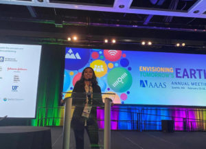 Young Woman presenting at a confrence. There is a large screen behind her. She is speaking into a microphone