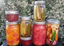 canned-food-in-jars.jpeg seven mason jars with tomatoes in one, pickle spears in two, red jam in two, a variety of vegetables in one jar with corn and other vegetables, and fruits in one jar.