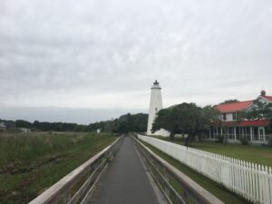 Wooden board walk leading to white light house with gray skies and tall grasses in the background.