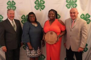 Four individuals pictured with award recipient in front of a green and white 4-H drop.