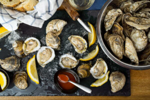 Dark marble serving platter with shucked oysters with sliced lemons and a sppon of cocktail sauce on a table next to a bucket of unshucked oysters and a basket with a tea towel and bread in it.