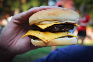 A hand holds a hamburger. With a lawn and people gathering in the background.