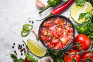 bowl of salsa surrounded by tomatoes, limes, peppers, and cilantros